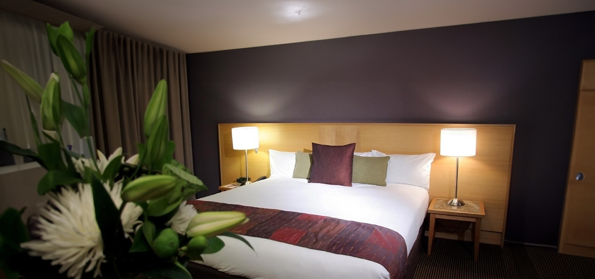 Modernes, sauberes Hotelzimmer mit einem großen Bett auf dem eine rot-weiße Decke liegt mit Pflanzen im Vordergrund