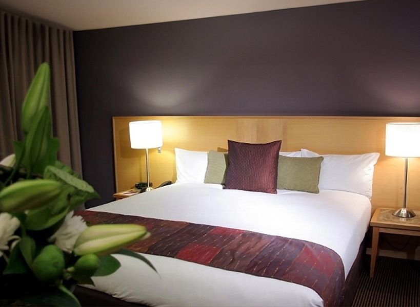 Modernes, sauberes Hotelzimmer mit einem großen Bett auf dem eine rot-weiße Decke liegt mit Pflanzen im Vordergrund