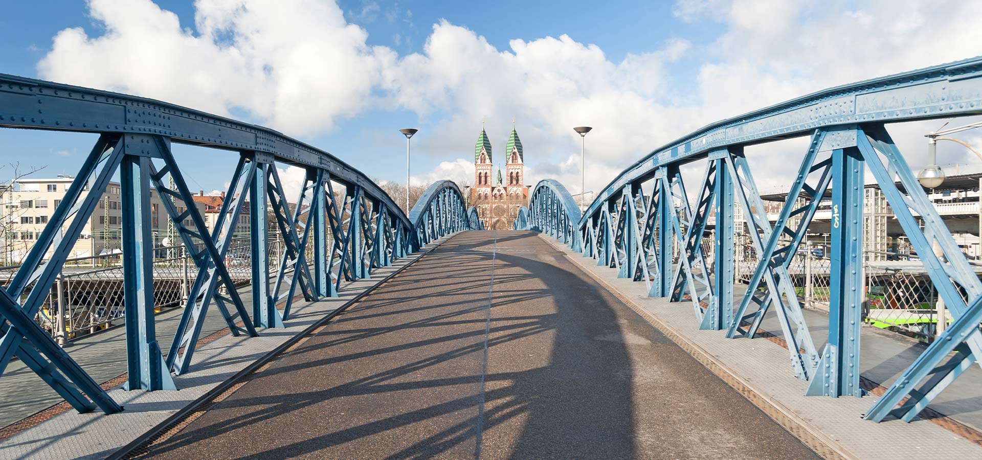 Blick auf leere, blaue Fachwerkbrücke die zu einem entfernten Gebäude mit zwei grünen Türmen führt