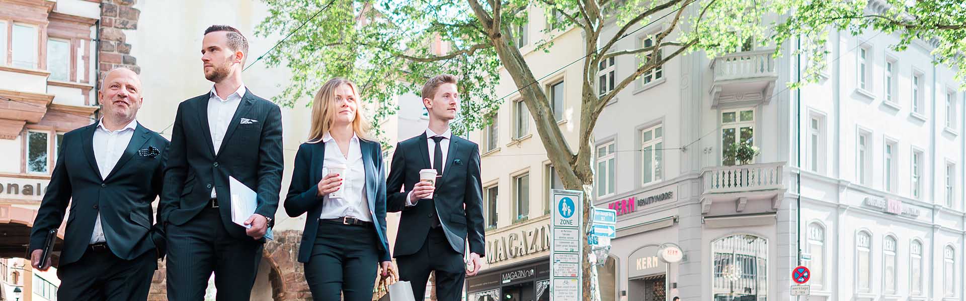 Vier Geschäftsleute in Anzügen halten Kaffees und Ordner und überqueren eine Stadtstraße vor einem Baum