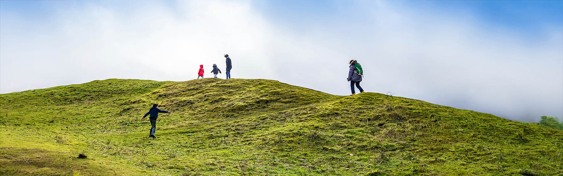Vier Wanderer laufen vor blauem Himmels mit tief hängenden Wolken über einen grasbewachsenen Hügel