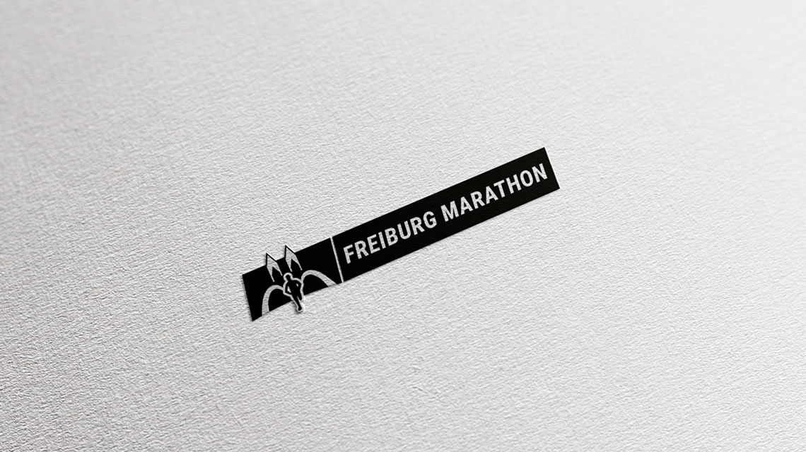 Ansicht des Logos von Freiburg Marathon neu in Schwarz auf Hellgrau und Papier