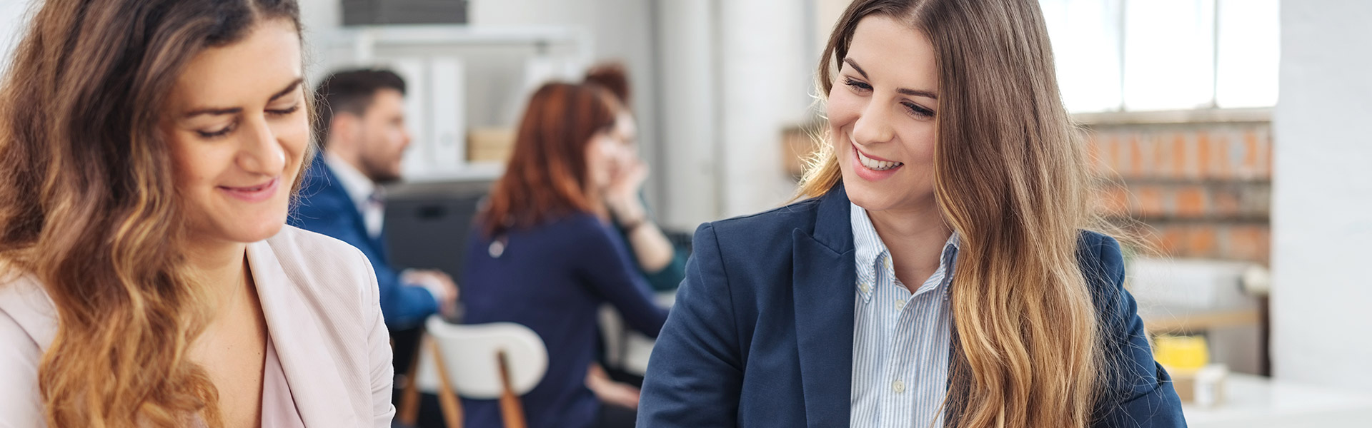 Zwei lächelnde Frauen sitzen an einem Schreibtisch in einem Büro und überprüfen Dokumente