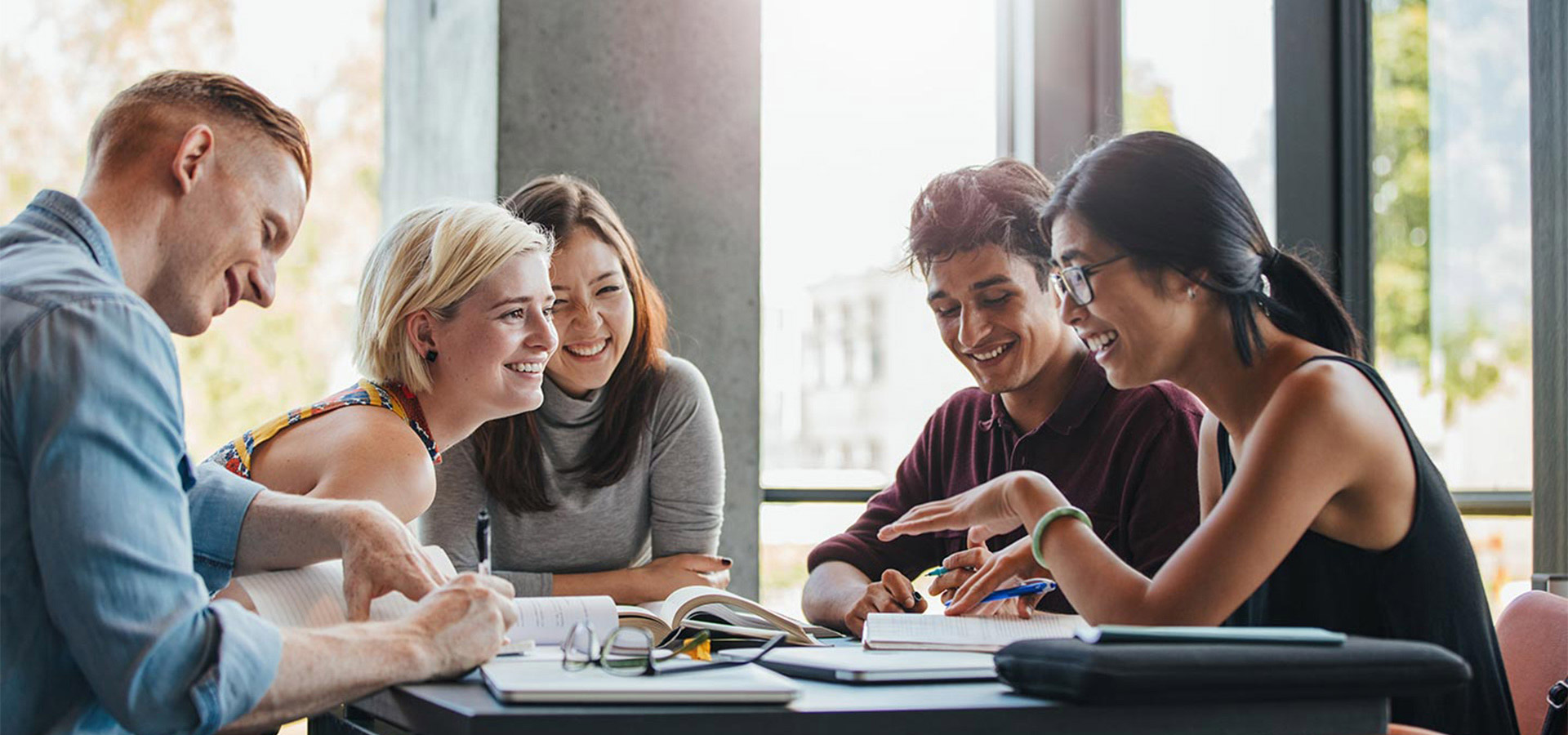 Gruppenbild mit drei Frauen und zwei Männer, die mit Büchern und Heften an einem Tisch lachend diskutieren