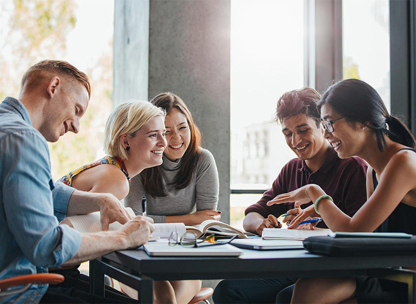 Gruppenbild mit drei Frauen und zwei Männer, die mit Büchern und Heften an einem Tisch lachend diskutieren